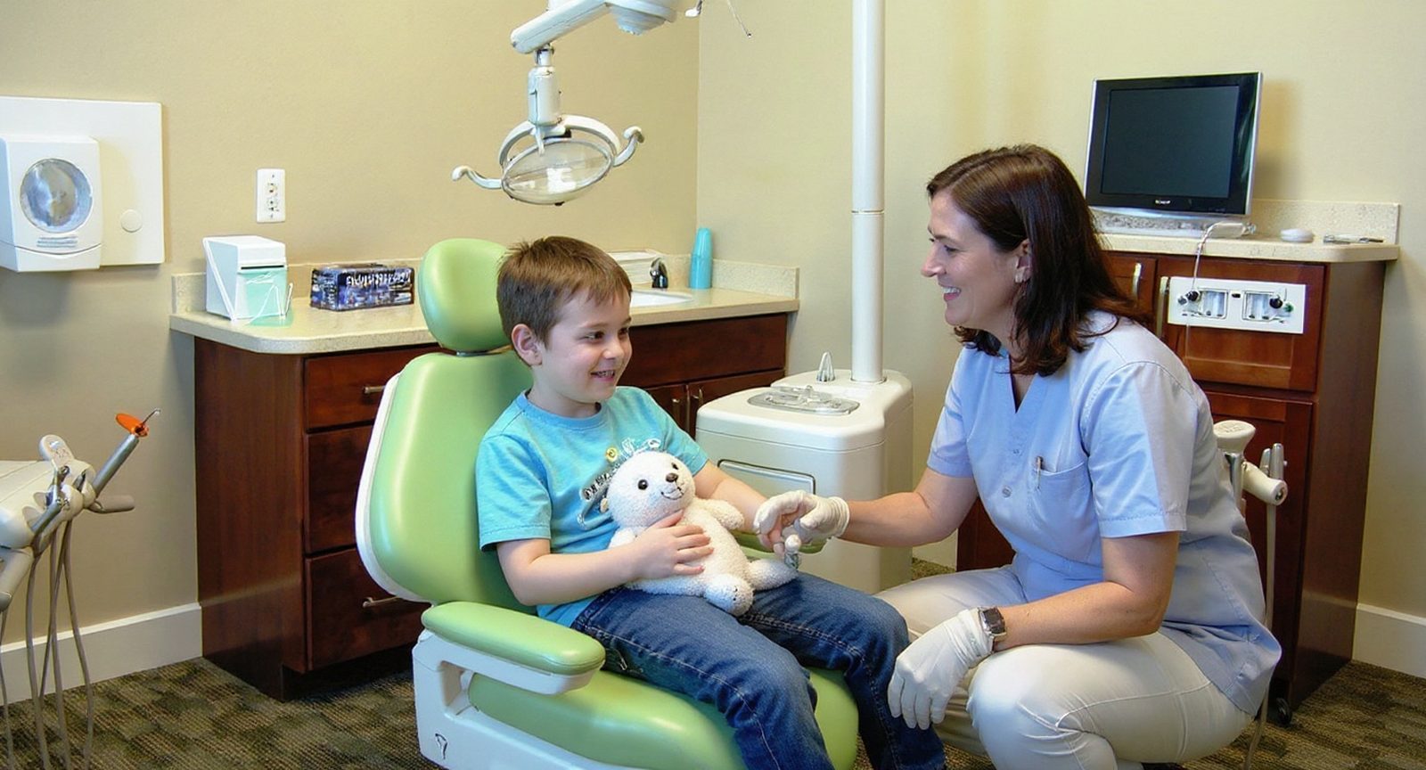 A pediatric dentist kneels to talk with a boy holding a stuffed animal in the dental chair, his mother beside him